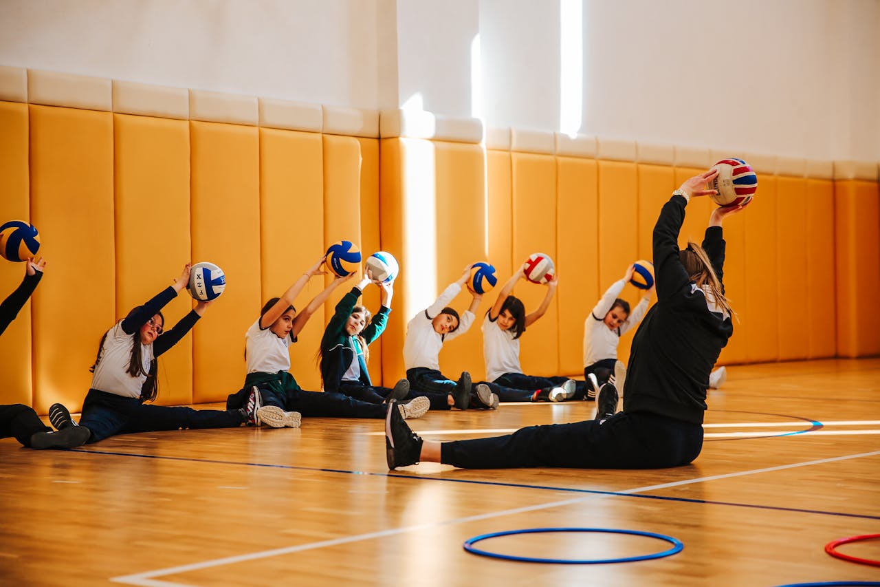 Group of children practicing volleyball stretches in a spacious indoor gym with a coach.